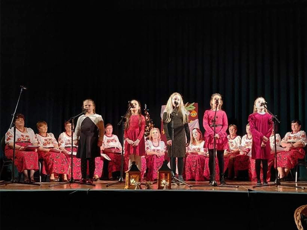 Na scenie na pierwszym planie stoi 5 dziewczynek, które śpiewa. Za nimi siedzą panie ubrane w ludowe biało-czerwone stroje. 